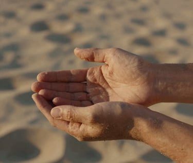 A cinematic close-up of two hands gently holding, captured in the warm golden hour light. The background features soft sand textures and muted tan tones typical of a Southern European coastal setting.