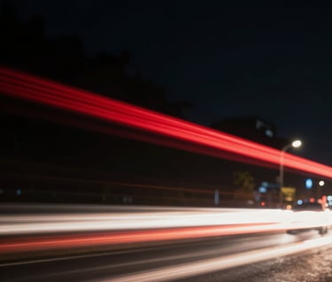 A blurred motion shot of city lights at night, dominated by deep black backgrounds and streaks of deep red and off-white light, suggesting speed and high production value.