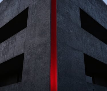 A dramatic low-angle shot of a minimalist modern building facade at night. The concrete is charcoal gray, with sharp geometric shadows in deep black. A single vertical strip of vivid deep red light runs down the center of the frame.