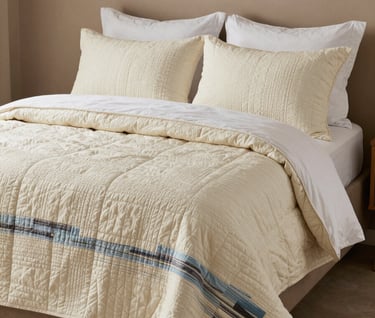 A lifestyle photograph of the Coastal Breeze quilt styled on a modern bed in a room with warm taupe walls and soft cream linens. The overall mood is peaceful and premium.