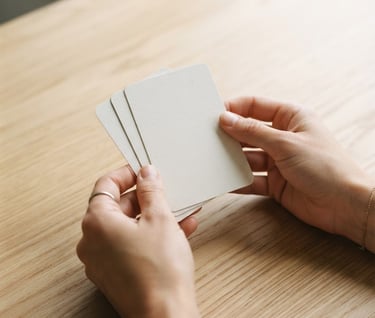 Close-up of hands holding clean therapy cards on a light wooden table, soft natural daylight, muted sophisticated tones