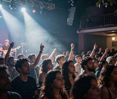 A candid, vibrant photograph of an audience at an intimate live music venue in a Latin American city. The stage lights create misty soft white and cool steel grey beams through the dark slate blue atmosphere. The mood is energetic and authentic.