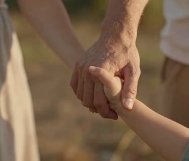 A close-up, cinematic shot of a mother and father's hands holding their toddler's small hand. The skin tones are warm and glowing under a late afternoon sun. The background is a soft-focus blur of Warm Brown garden elements.