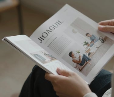 Close-up spontaneous shot of a person's hand holding a high-end lifestyle magazine, turning a page. Cinematic lighting, soft focus on the background. Colors include #9A9E96 and #F8F6F2. Elegant and professional style.