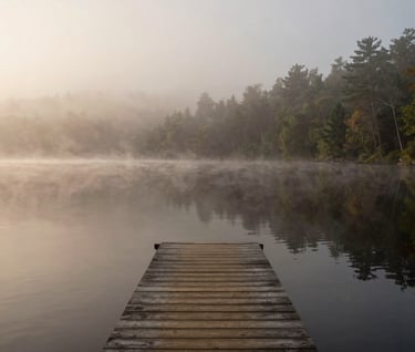 A serene, misty morning on a calm lake in a North American / US national park. A small wooden dock leads the eye into the soft off-white fog. The water is a dark, glassy charcoal. Warm, inviting, and quiet atmosphere.