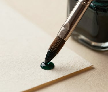 Macro photograph of a delicate paintbrush touching a piece of heavy-grain cotton paper. A small droplet of dark forest green ink is about to spread. The background is a blurred warm cream.
