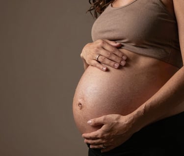 An emotional shot of a pregnant woman's hands gently resting on her belly, wearing a ring that catches the soft studio light. The setting is a South American / Brazilian professional studio with a minimalist aesthetic and earthy brown tones.