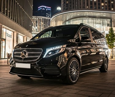 Black Mercedes-Benz V-Class luxury passenger van parked on a city street at night with glowing city lights.