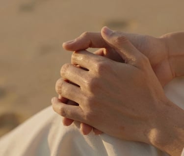 Extreme close-up of a couple's hands gently intertwined. The lighting is warm and cinematic, reflecting a golden oak glow. Soft focus on a background featuring warm sand tones and soft off-white fabrics.