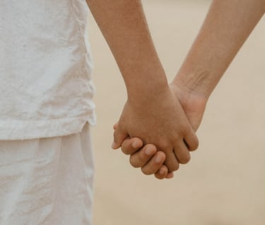 A close-up, rectangular photograph of a child's hand holding a parent's hand. The scene is shot in a warm, sun-drenched environment with a cinematic, soft-focus background. The tones are natural, featuring white linen textures and soft sand-colored skin tones. Authentic and emotional.