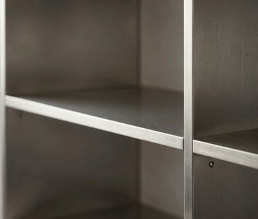 Side view of a luxury stainless steel bookshelf. The metal is polished to a mirror finish, reflecting a minimalist interior. Technical precision in every corner and joint.