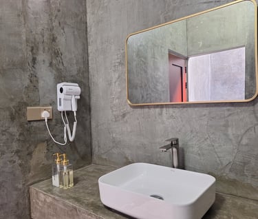 Modern industrial bathroom with a white vessel sink, gold framed mirror, and hair dryer against concrete walls.