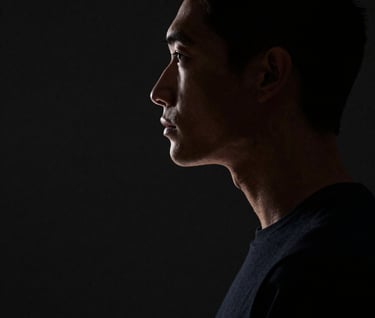 A profile view photograph of a person in a minimalist studio. Only the edges of the face and torso are caught in a thin sliver of off-white light, with the rest of the frame immersed in dark black negative space. North American / US.