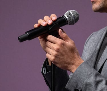A close-up artistic shot of Stefan Taylor's hands on a microphone stand. The lighting is soft lavender grey against a muted plum background, highlighting the sophisticated and professional polish of his performance attire.