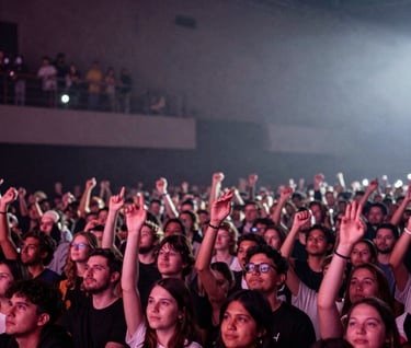 A wide-angle shot of a cheering crowd in a dimly lit venue, seen from the stage perspective. Hints of deep mauve and misty white lights illuminate the fans. Dramatic and engaging concert atmosphere.