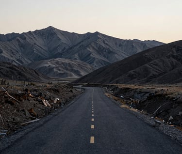 A breathtaking landscape of a winding asphalt road cutting through a vast, desolate Global / Western mountain range. The sky is a soft off-white, contrasting with the deep slate grey of the peaks and the rich charcoal black of the road surface.