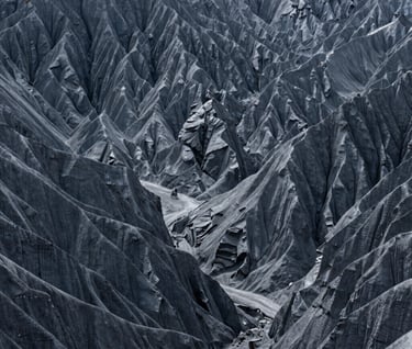 A sprawling, minimalist landscape of Global / Western canyon walls. The rock faces are shades of dark grey and cool grey. The composition is dominated by negative space, with a small, distant motorcycle visible for scale.
