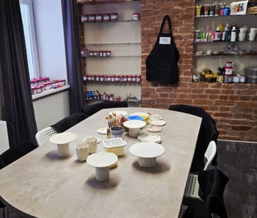 Pottery studio workshop table with unpainted ceramic clay pieces, brushes, and paints against a rustic brick wall.