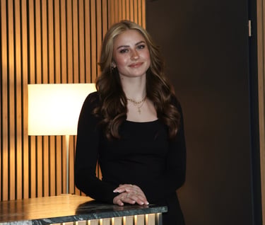 Smiling young woman with wavy blonde hair in a black dress posing by a marble counter.