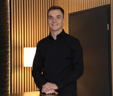 Smiling young businessman in a black button-down shirt posing in a modern office with wood paneling.