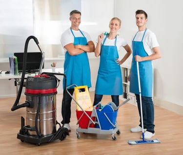 Three smiling people wearing blue aprons ready to clean with mops, cleaning bucket, supplies and an industrial vacuum