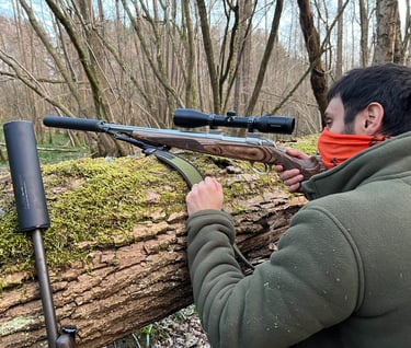 A stalker in a green fleece taking aim with a scoped rifle moderator rested on a moss-covered fallen