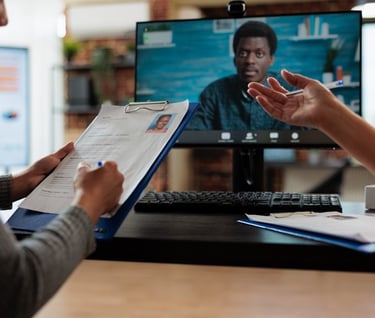 A figure looks distractedly at a CV on a clipboard whilst an interview candidate appears on screen