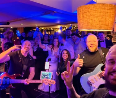 A live wedding band with guitars posing for a selfie with a cheering crowd on a blue lit dance floor.