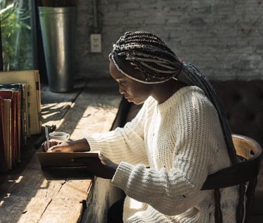 Young woman writing in a journal at a rustic wooden desk by a sunny window with books.