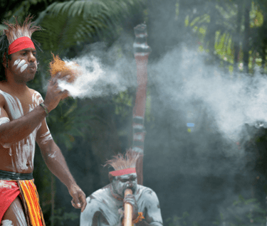 aborignal men one playing a didgeridoo and 1 doing a smoking ceremony