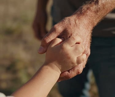 A tight, candid close-up of a father's hands holding his young child's hands. The lighting is warm and sun-drenched, with terracotta highlights on their skin. Set in an outdoor North American / General Western environment, the focus is on the emotional connection.