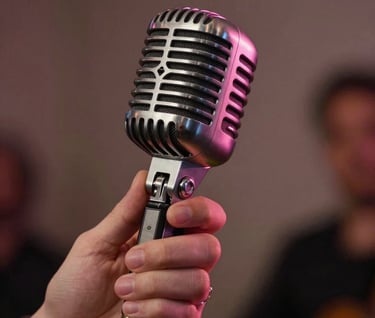 Close-up photography of a performer's hands holding a vintage-style microphone in a North American / US jazz club. Soft pink lighting accents the metallic textures, creating a mood of modern professionalism and timeless grace.