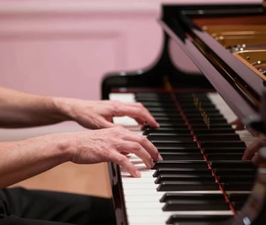 Elegant hands of a musician playing a grand piano in a prestigious North American / US concert hall. The lighting is a gentle mist of soft pink, highlighting the polished wood and the delicate, graceful movement of the fingers.