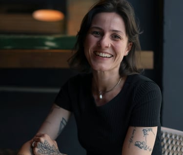Smiling woman with tattoos sitting at a cafe table, wearing a black shirt and white pants.