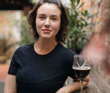 A woman in a black shirt sits at a rustic outdoor bar with a glass of dark craft beer.