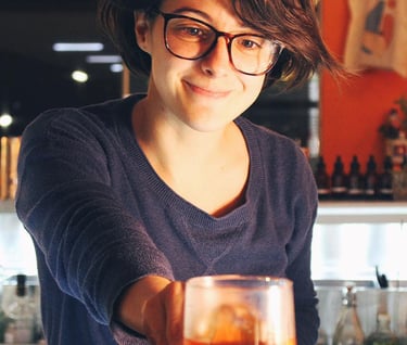 Smiling female bartender serving a craft cocktail in a glass at a dimly lit bar counter.