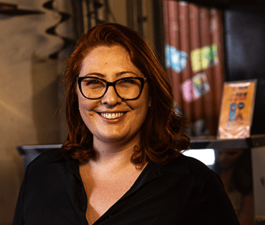 Smiling woman with red hair and black glasses wearing a dark button-down shirt in a cafe.