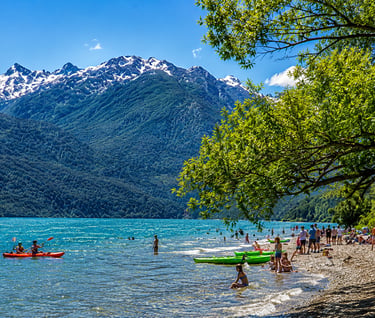 Tourists kayaking and swimming at a sunny mountain lake beach with snow-capped peaks in the background.