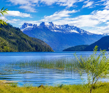 Snow-capped mountain range reflecting in a clear blue lake surrounded by lush green forests.