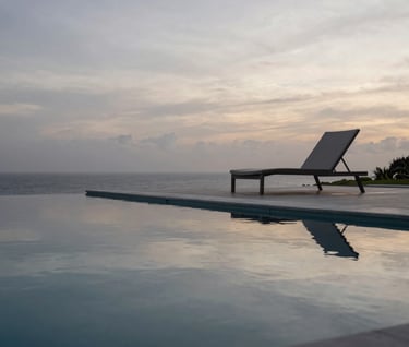 A detail shot of a black infinity pool reflecting the silver grey sky at dawn. A single minimalist lounge chair sits on the edge. The setting is a private estate in a South American / Brazilian coastal region. Sophisticated and exclusive atmosphere.