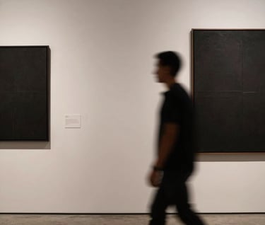 Photography of a blurred human silhouette moving through a minimalist South American / Argentine art gallery. The composition plays with shadows against a soft off-white wall, with accents of deep charcoal black.