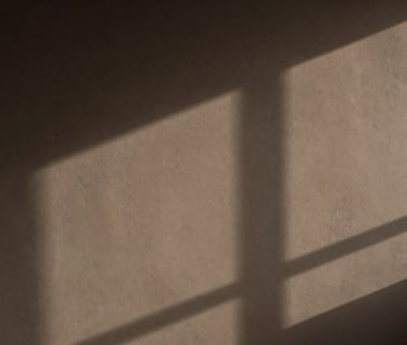 A minimalist, contemplative photograph of a shadow cast on a wall in a South American / Argentine studio. The wall has a subtle texture in earthy taupe, and the interplay of light and shadow suggests a deeper, enigmatic presence behind the material surface.