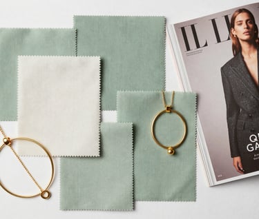 Top-down editorial photography of a curated style board with fabric swatches in soft sage green and soft off-white, paired with gold jewelry pieces and a high-end fashion magazine, set in a North American / Western European studio.