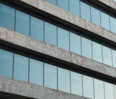 A detailed shot of modern building textures, combining raw concrete in light gray with large panes of glass reflecting a clear baby blue sky, South American urban architecture, minimalist and clean composition.