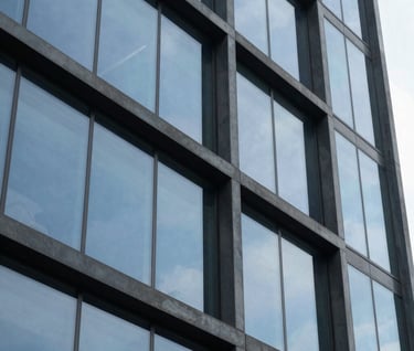A close-up photograph of architectural glass reflecting a baby blue sky and dark grey steel structures. The lighting is crisp, highlighting the textures and clean lines of modern South American design.
