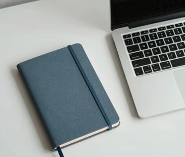 A minimalist overhead shot of a professional workspace in a Southeast Asian / Indonesian setting, featuring a clean desk, a blue-grey notebook, and a sleek laptop, emphasizing quiet competence.