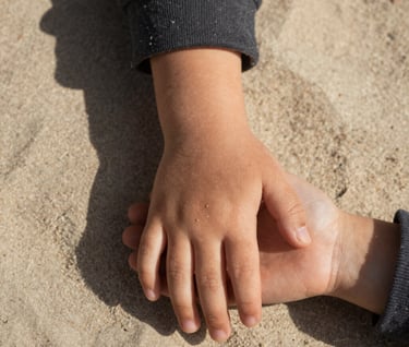 Macro shot of a child's hand holding a parent's weathered hand against a soft sand colored linen background. Set in a North American / US garden, warm afternoon light creating deep, inviting shadows. Charcoal tones in the clothing.