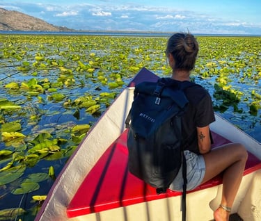 A traveler wearing a black waterproof backpack sits on a boat in a lake covered in water lilies.