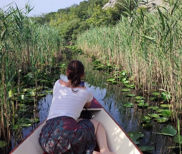 A woman sits in a boat on a narrow river path surrounded by tall reeds and lily pads.