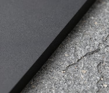 Macro photography of an industrial material, showing the transition between a smooth charcoal surface and a rough stone gray texture. The composition is abstract and editorial, shot with a shallow depth of field in a studio setting.